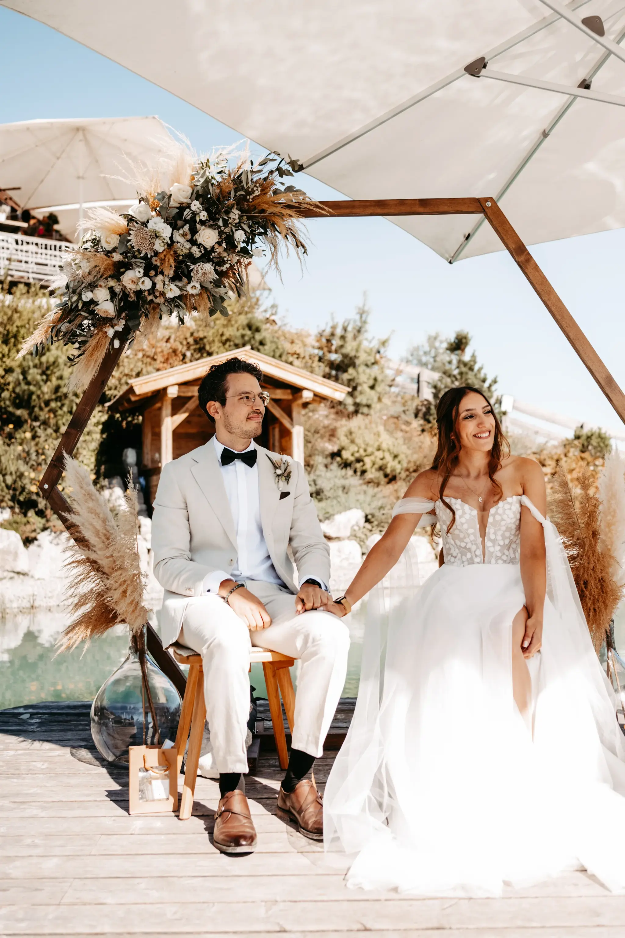 Bride and groom during their open-air ceremony under a parasol on the deck by the Stöttlalmsee, with a floral arch in Tyrol