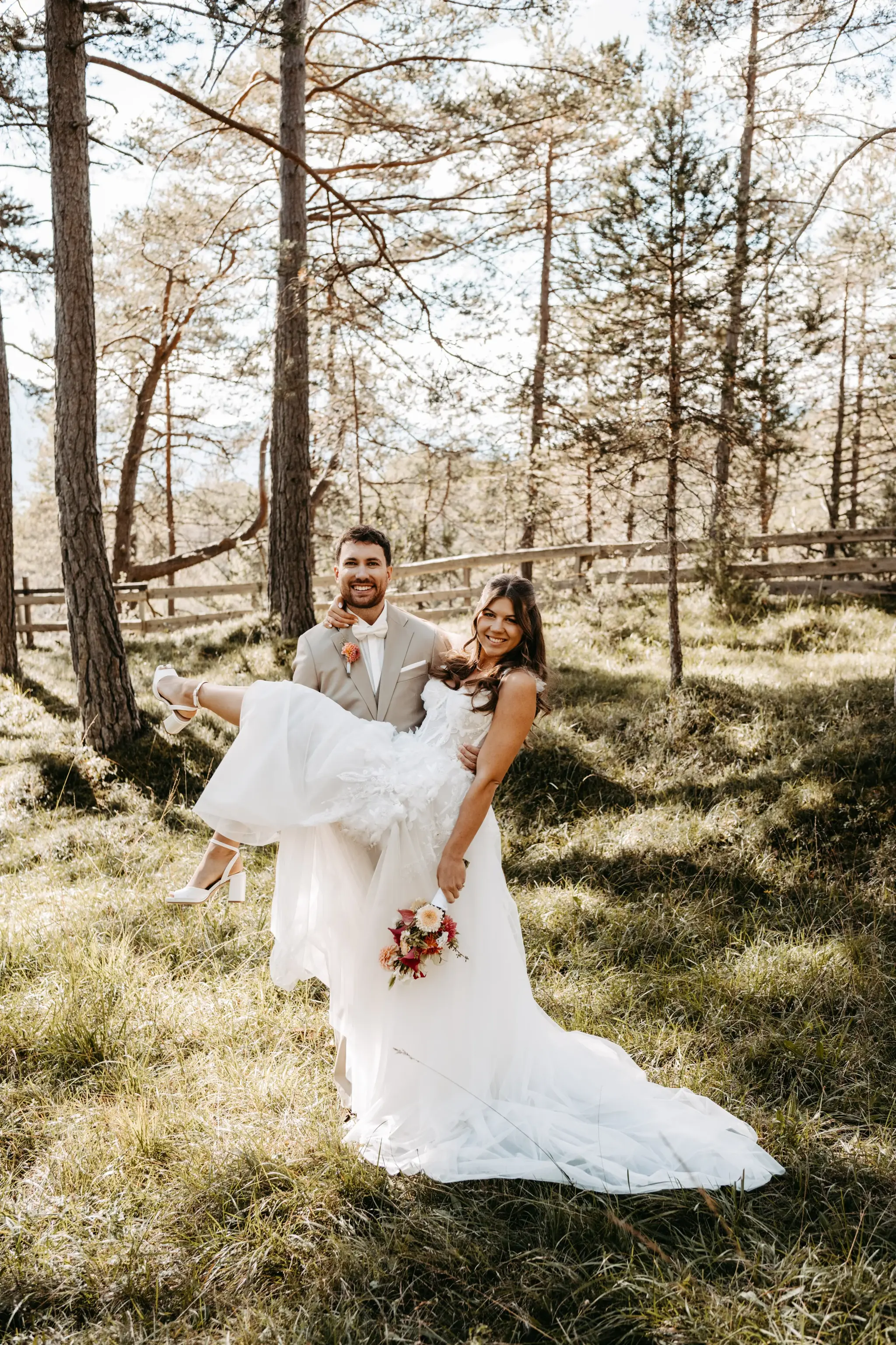 Smiling groom carrying his bride through a sunlit pine forest near the Stöttlalm in Mieming, Tyrol