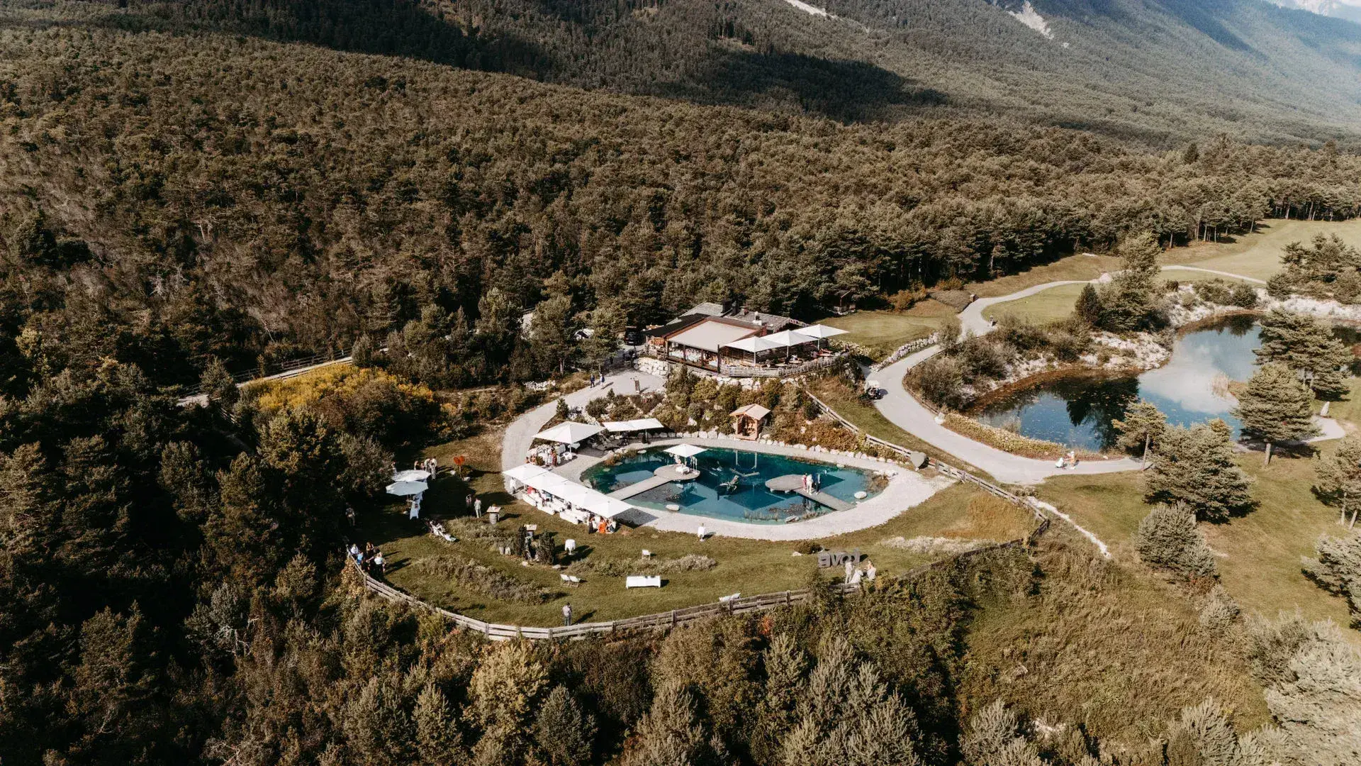 Aerial view of the Stöttlalm with its turquoise bathing pond and pavilions nestled in forest on the Mieminger Plateau