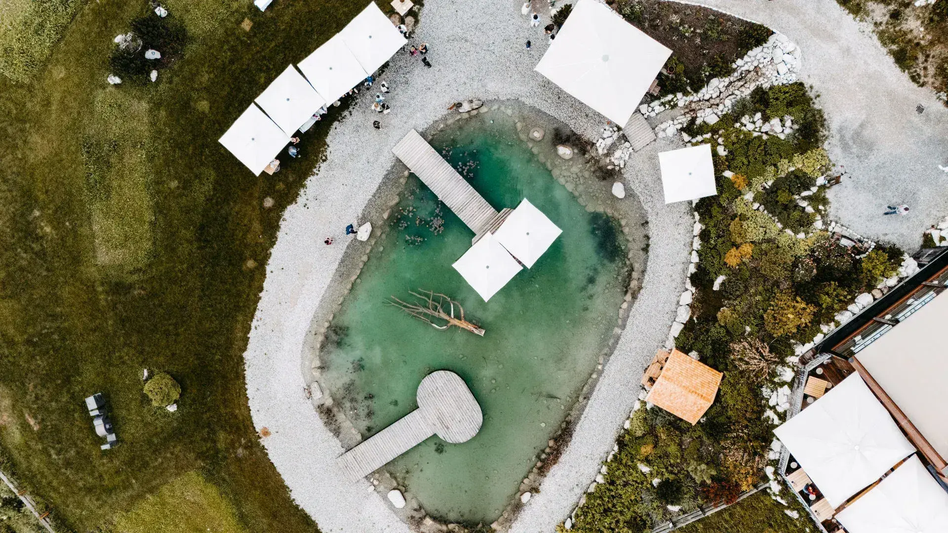 Overhead aerial view of the turquoise Stöttlalmsee with wooden jetty and white event pavilions amid meadows in Tyrol
