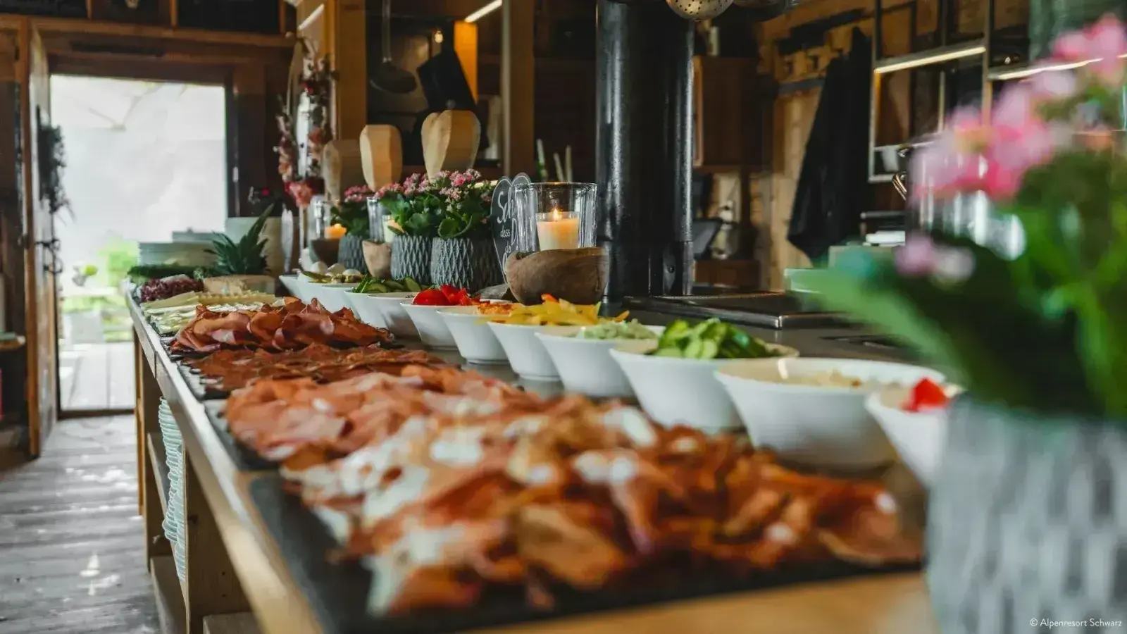 Langes Frühstücksbuffet mit Aufschnitt, Schinken, Gemüse und Salaten, brennender Kerze und Blumenschmuck in der gemütlichen Almstube der Stöttlalm
