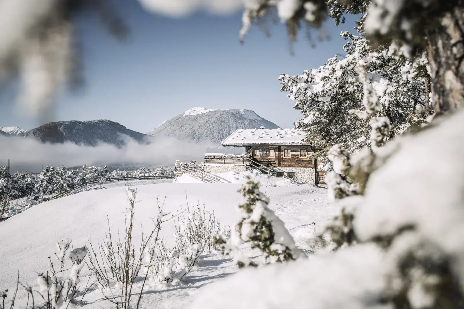 Tief verschneite Stöttlalm in der Wintersonne zwischen verschneiten Kiefern, dahinter Berge und Nebel über dem Tal